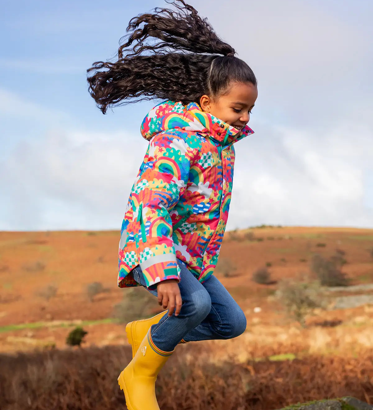 Child jumping and wearing the Frugi snow and ski coat in the fun frenzy design and yellow wellington boots in a grassy field