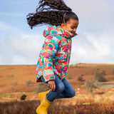 Child jumping and wearing the Frugi snow and ski coat in the fun frenzy design and yellow wellington boots in a grassy field