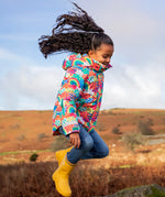 Child jumping and wearing the Frugi snow and ski coat in the fun frenzy design and yellow wellington boots in a grassy field