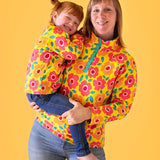A mother holding her daughter in front of a light orange background and wearing the yellow Frugi organic cotton fleece with a repeating floral print and blue jeans