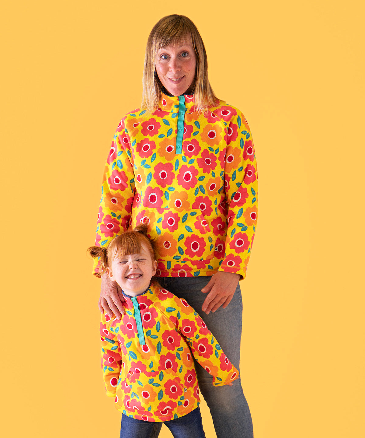 A mother standing behind her daughter with a light orange background. Both wearing the yellow Frugi organic cotton fleece with a repeating floral print and blue jeans