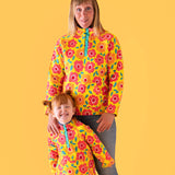 A mother standing behind her daughter with a light orange background. Both wearing the yellow Frugi organic cotton fleece with a repeating floral print and blue jeans