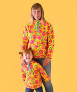 A mother standing behind her daughter with a light orange background. Both wearing the yellow Frugi organic cotton fleece with a repeating floral print and blue jeans