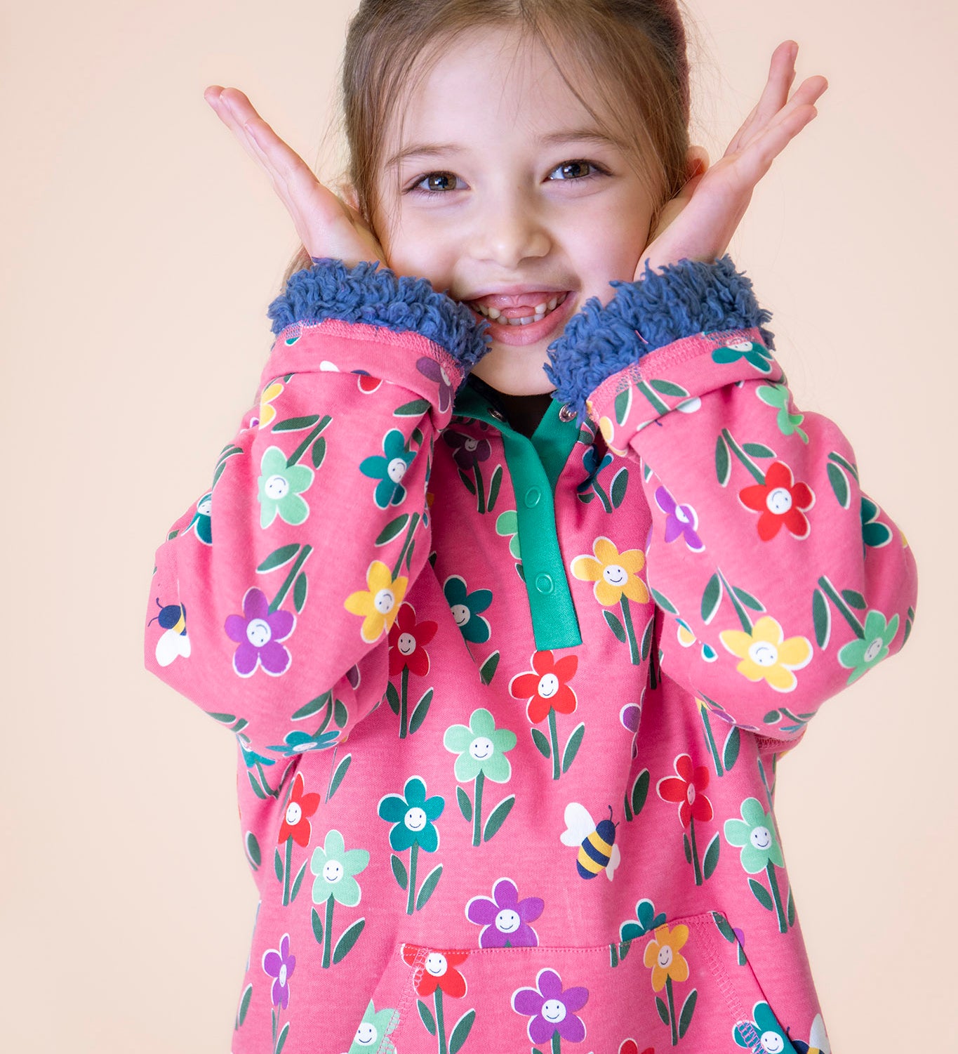 A child wearing the Frugi fun flowers snuggle fleece available at Babipur. A GOTS organic cotton thick jacket in pink with a playful flowery design and a purple fleece inner. Features a popper fastener in green trim