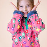 A child wearing the Frugi fun flowers snuggle fleece available at Babipur. A GOTS organic cotton thick jacket in pink with a playful flowery design and a purple fleece inner. Features a popper fastener in green trim
