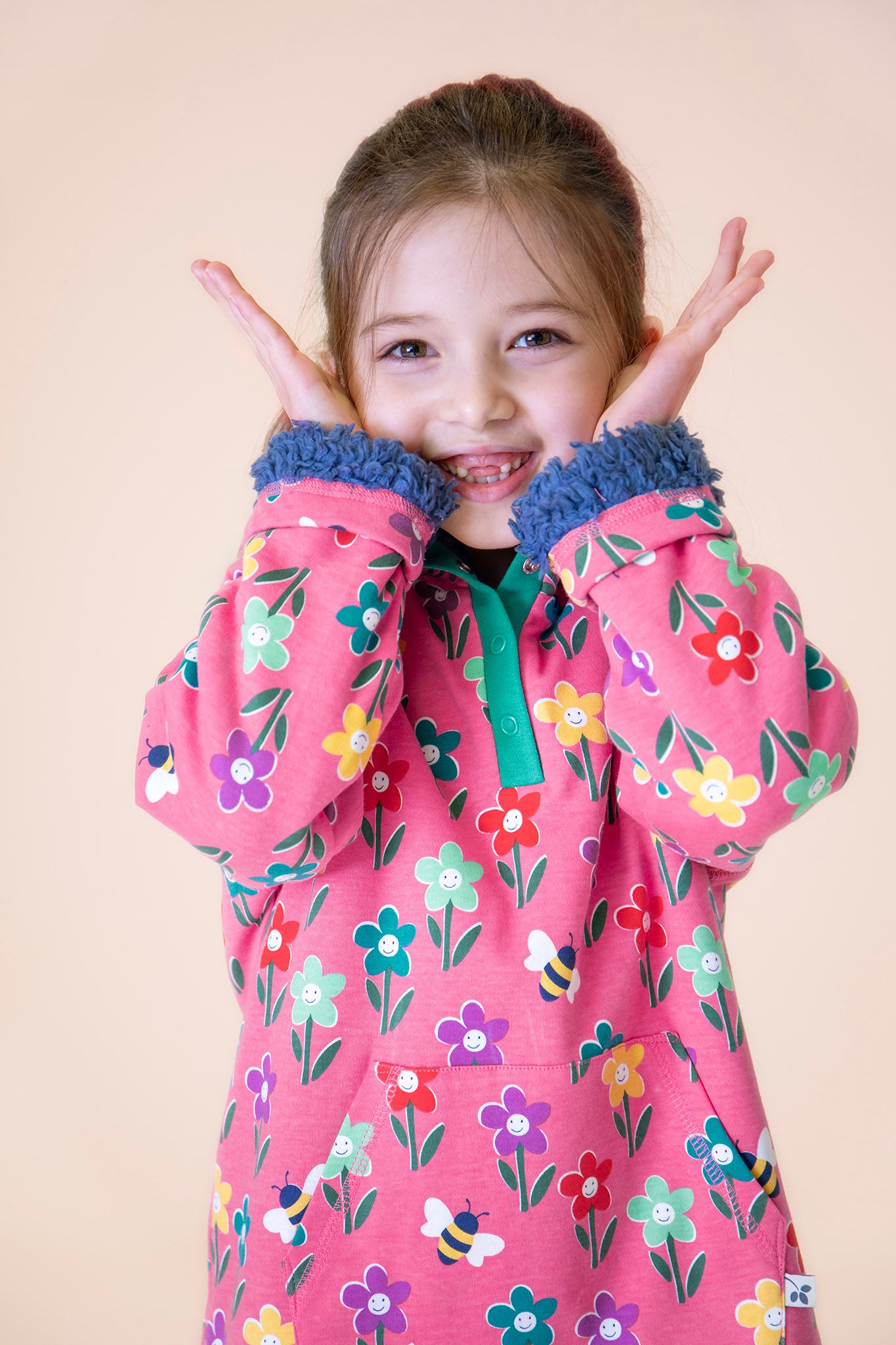 A child wearing the Frugi fun flowers snuggle fleece available at Babipur. A GOTS organic cotton thick jacket in pink with a playful flowery design and a purple fleece inner. Features a popper fastener in green trim