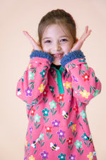 A child wearing the Frugi fun flowers snuggle fleece available at Babipur. A GOTS organic cotton thick jacket in pink with a playful flowery design and a purple fleece inner. Features a popper fastener in green trim