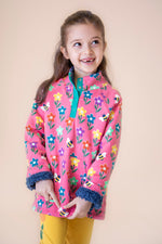 A child wearing the Frugi fun flowers snuggle fleece available at Babipur. A  thick pink jacket made from GOTS organic cotton with a playful flowery design and a purple fleece inner. Features a popper fastener in green trim