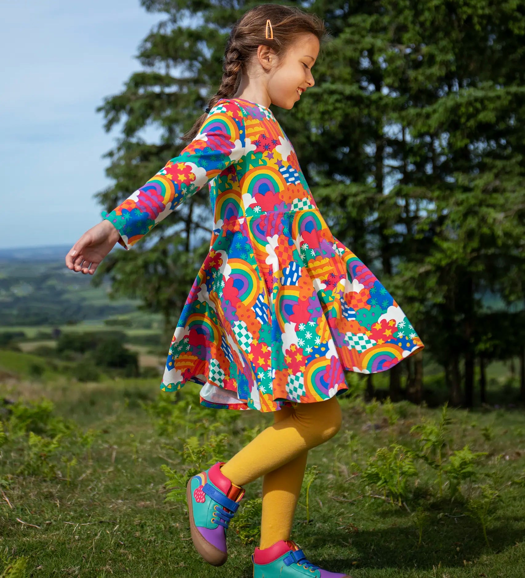 Frugi sofia skater dress in rainbow fun frenzy print, knee length with long sleeves, worn by child playing outside.