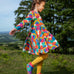 Frugi sofia skater dress in rainbow fun frenzy print, knee length with long sleeves, worn by child playing outside.