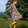 Frugi sofia skater dress in rainbow fun frenzy print, knee length with long sleeves, worn by child playing outside.