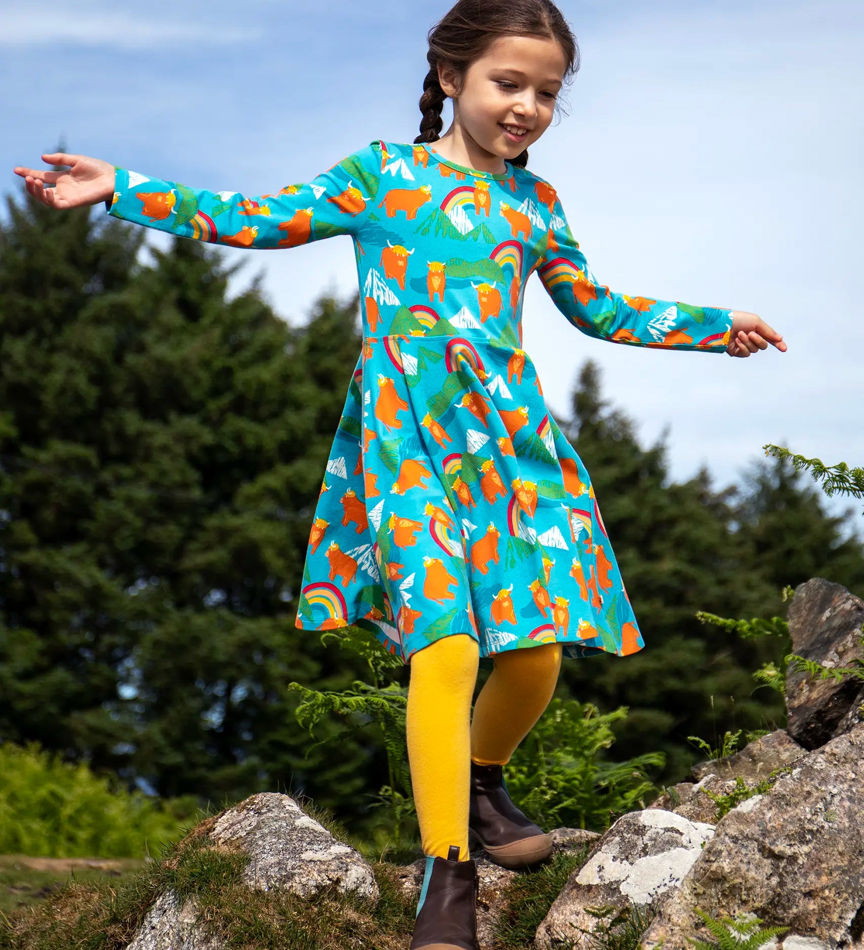 Frugi sofia skater dress in blue Highland cow print, knee length with long sleeves, worn by girl outside.