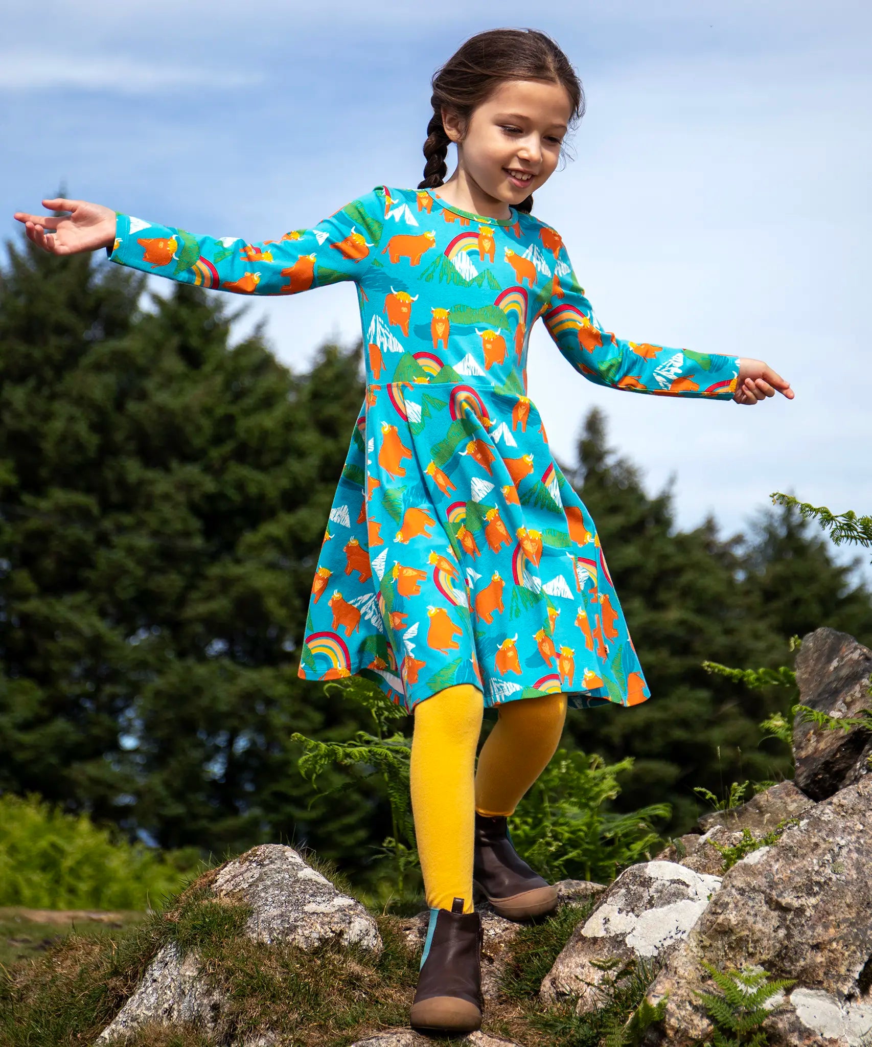 Frugi sofia skater dress in blue Highland cow print, knee length with long sleeves, worn by girl outside.