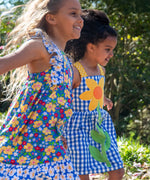 Child wearing Frugi's blue and white check dress with large sunflower patch on front next to child wearing a floral dress