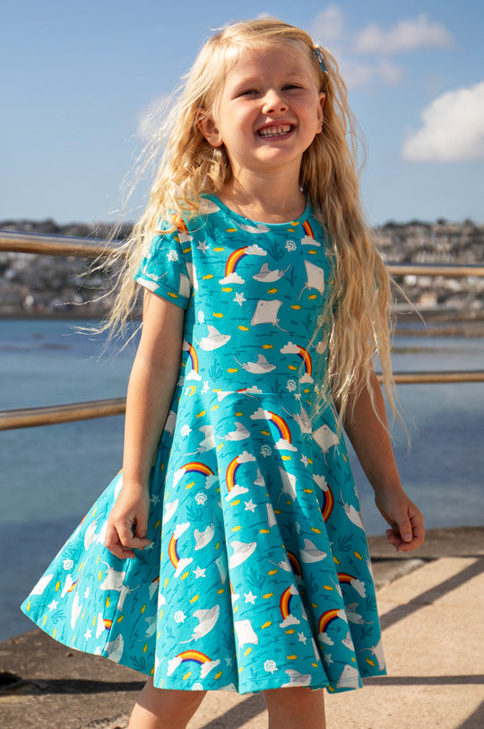 Frugi Spring organic cotton Skater Dress in Rainbow and sting Ray all-over print on blue background, worn by child on a harbour to represent new Frugi at Babipur.