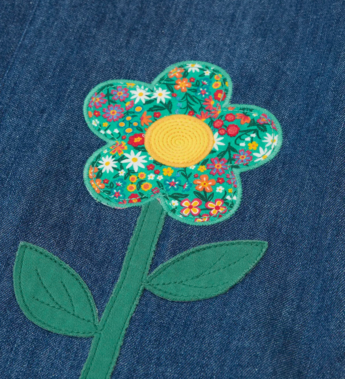 Frugi mid wash denim kids pinafore dress laid flat showing a close up of a decorative flower applique on the gather on organic cotton fabric