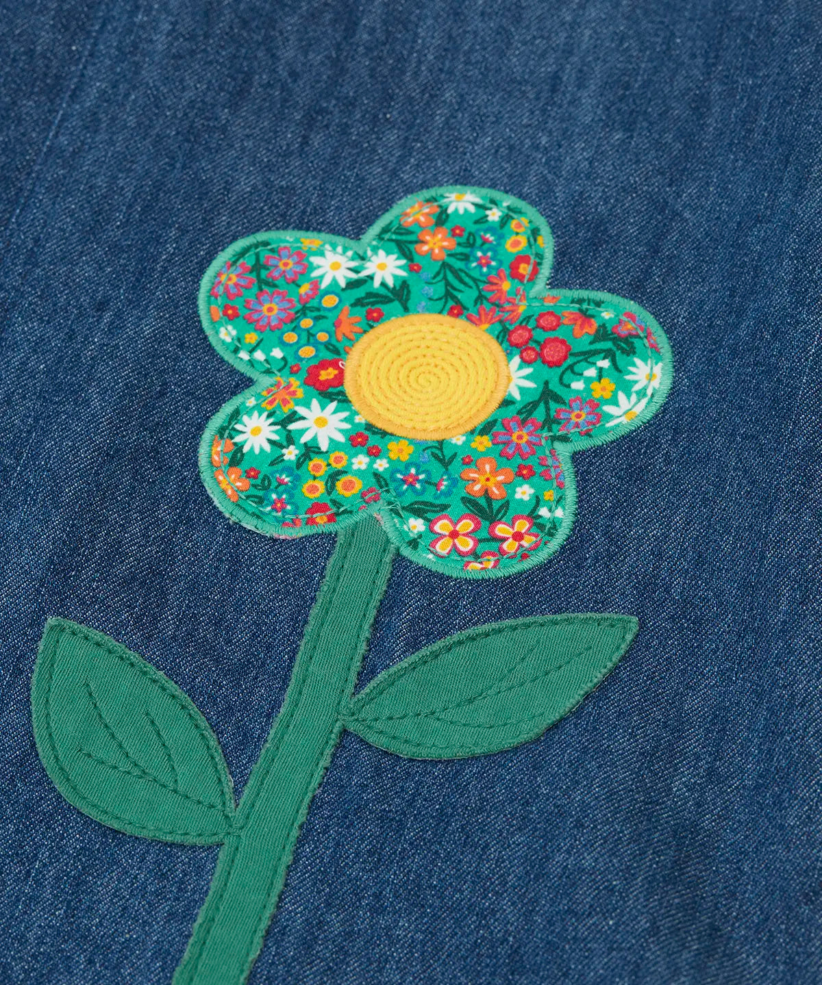 Frugi mid wash denim kids pinafore dress laid flat showing a close up of a decorative flower applique on the gather on organic cotton fabric