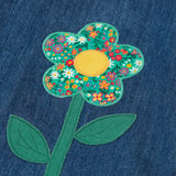 Frugi mid wash denim kids pinafore dress laid flat showing a close up of a decorative flower applique on the gather on organic cotton fabric