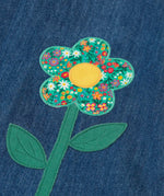 Frugi mid wash denim kids pinafore dress laid flat showing a close up of a decorative flower applique on the gather on organic cotton fabric