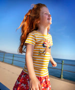 Child wearing Frugi's yellow and white stripe short sleeve t-shirt with a horse patch on the front and standing on the promenade