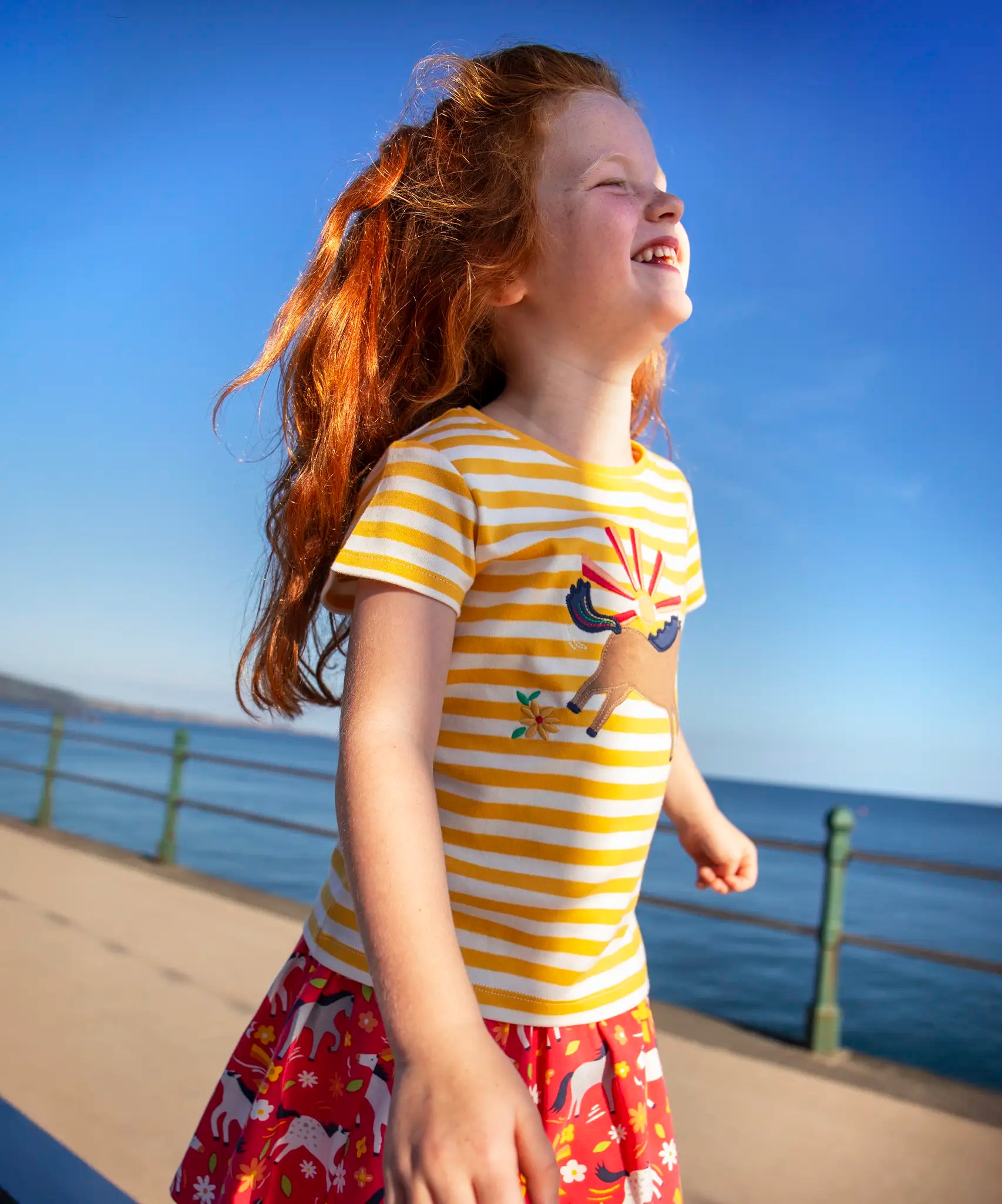 Child wearing Frugi's yellow and white stripe short sleeve t-shirt with a horse patch on the front and standing on the promenade