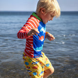Child wearing Frugi's red and white striped sun safe rash vest and blue body with different fish and standing in the ocean