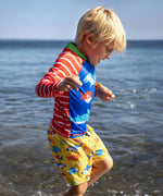 Child wearing Frugi's red and white striped sun safe rash vest and blue body with different fish and standing in the ocean