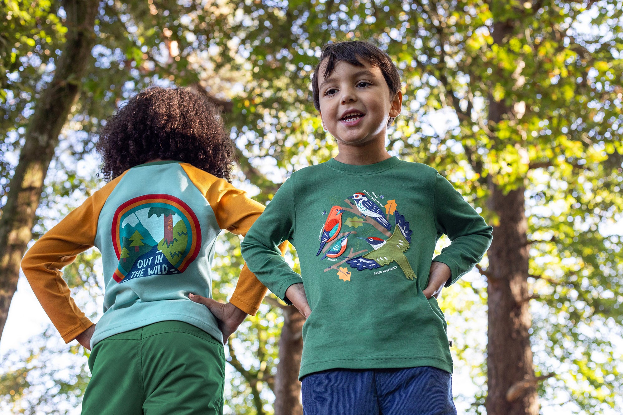 A child standing outside by trees and wearing the Frugi kids terrific applique top in holly green/birds available at Babipur.