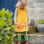 A child stood outside a building and looking down. The child is wearing the Frugi Lovely Lily Cord Dress - Gold/Red Panda, and the Frugi Norah Tights - Red Panda Pals, along with a Frugi rainbow stripe top underneath the dress.