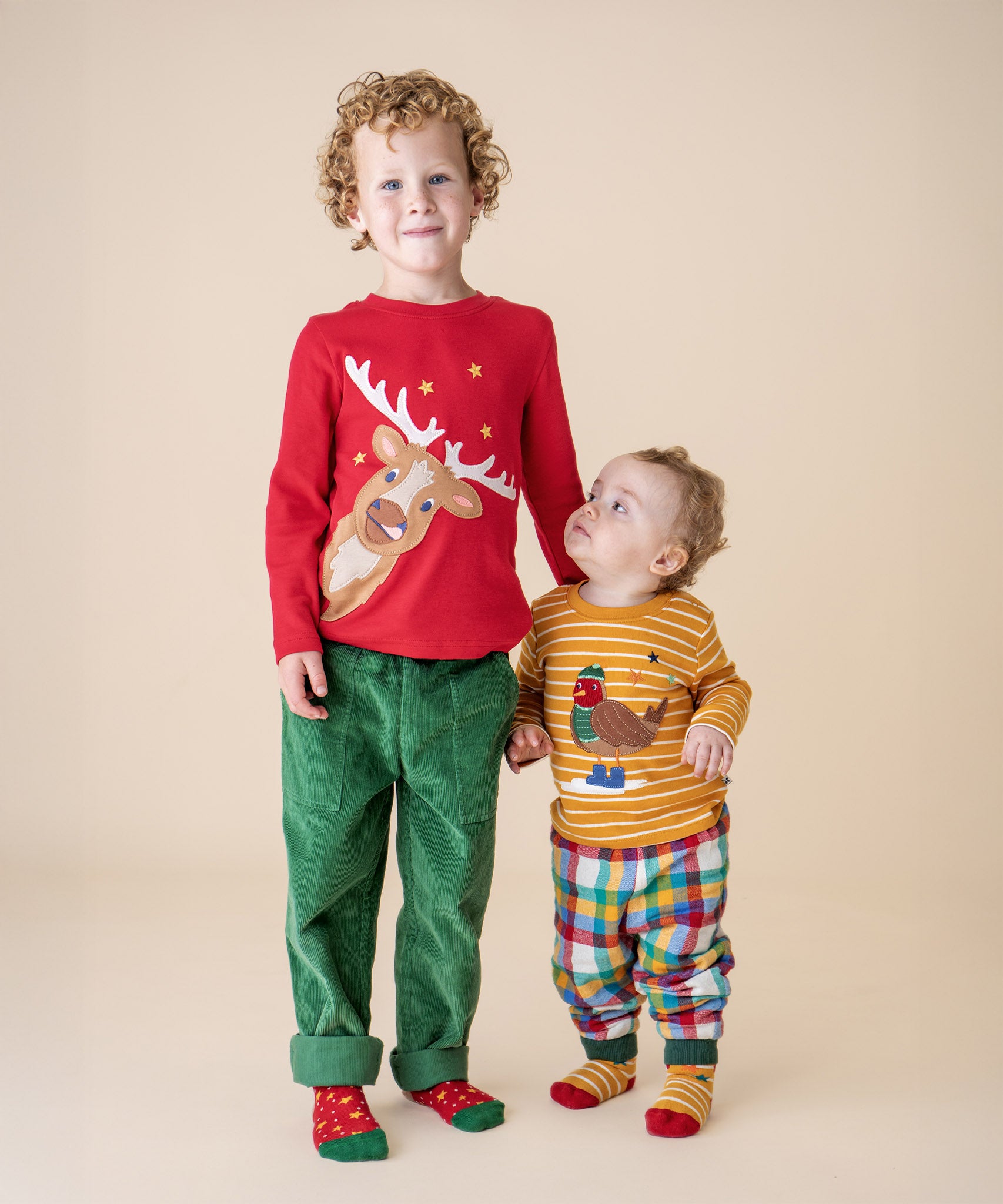 A boy wearing the Frugi Berry Red Reindeer Terrific Circular Organic Cotton long sleeved kids top standing next to a small child wearing the gold stripe robin appliqué top. These tops are both available at Babipur. 