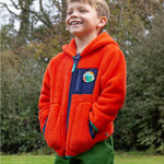 A child outside wearing the Frugi Toby Ted Fleece Jacket - Bonfire, with green Frugi trousers