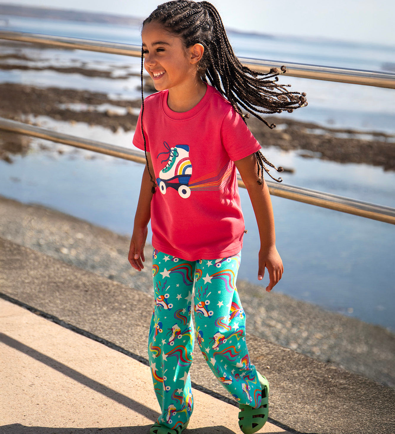 Toddler standing on a concrete surface with a beach in the background. Wearing the light pink Frugi short sleeve top with a roller blade design and turquoise trousers