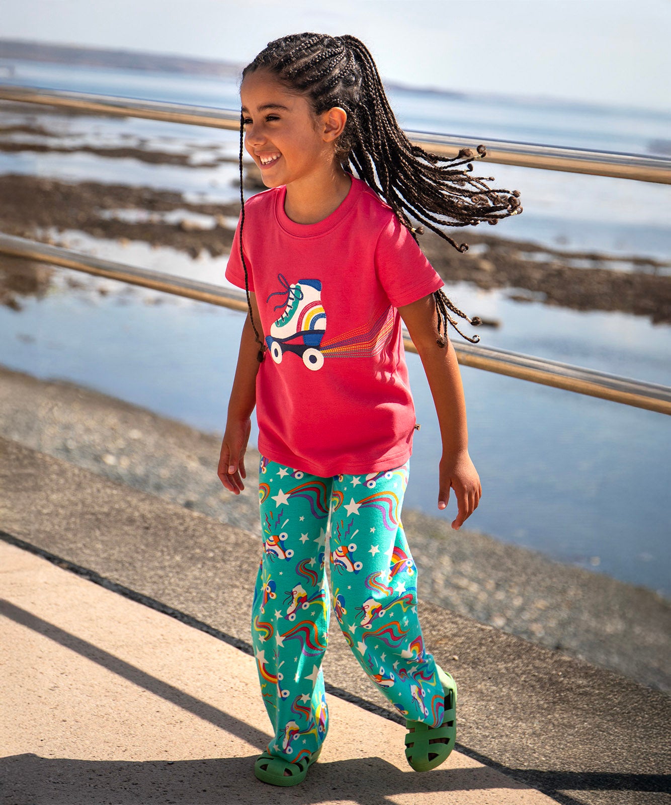 Toddler standing on a concrete surface with a beach in the background. Wearing the light pink Frugi short sleeve top with a roller blade design and turquoise trousers