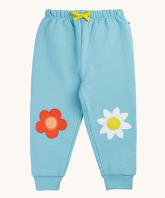 Frugi organic cotton joggers laid flat. In light blue with a red flower applique on one knee and a white flower on the other knee. Features a yellow tie on the waist band