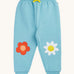 Frugi organic cotton joggers laid flat. In light blue with a red flower applique on one knee and a white flower on the other knee. Features a yellow tie on the waist band