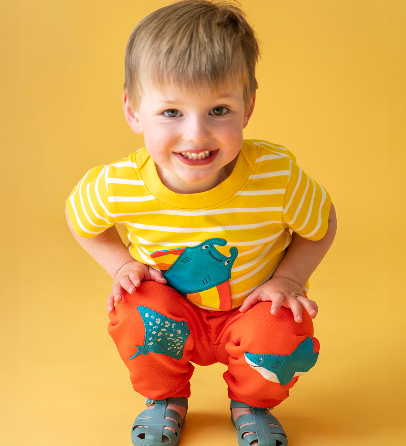 Child smiling and crouching in front of a light orange background. Wearing a yellow and white striped t-shirt and the bright orange joggers with a manta ray patch on one knee and a blue shark on the other knee