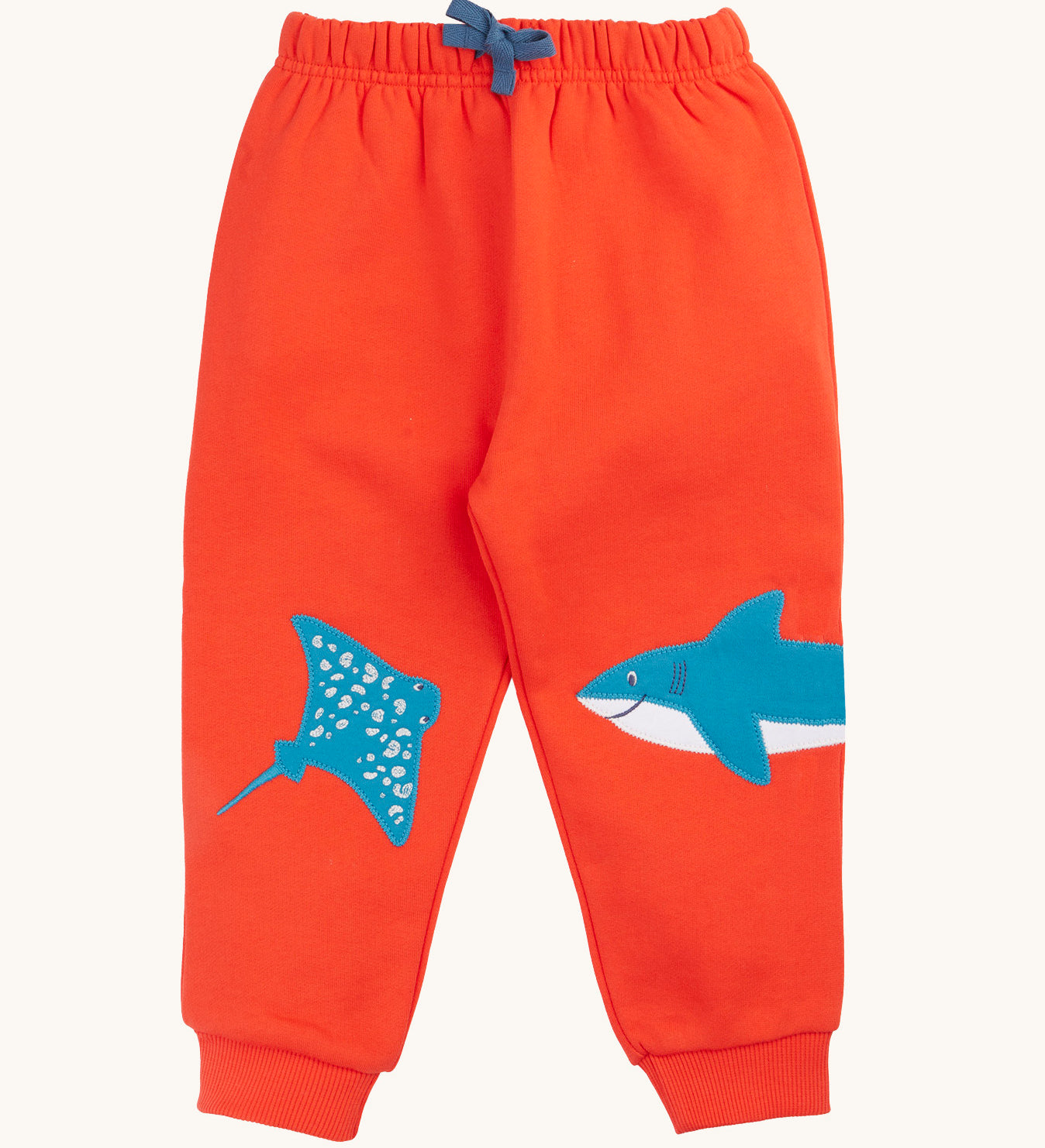 Frugi organic cotton joggers laid flat. In bright orange with a blue manta ray patch on one knee and a blue shark on the other knee. Features an elasticated waist band with blue tie