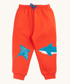 Frugi organic cotton joggers laid flat. In bright orange with a blue manta ray patch on one knee and a blue shark on the other knee. Features an elasticated waist band with blue tie
