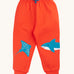 Frugi organic cotton joggers laid flat. In bright orange with a blue manta ray patch on one knee and a blue shark on the other knee. Features an elasticated waist band with blue tie