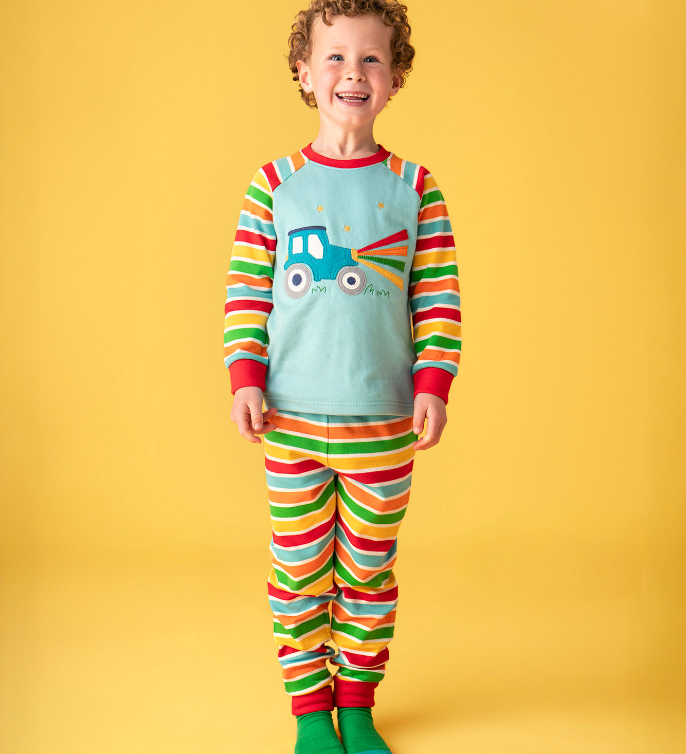 A child smiling and standing in front of a light orange background. Wearing the light blue pyjama top with striped sleeves and a tractor on the front and matching striped trousers