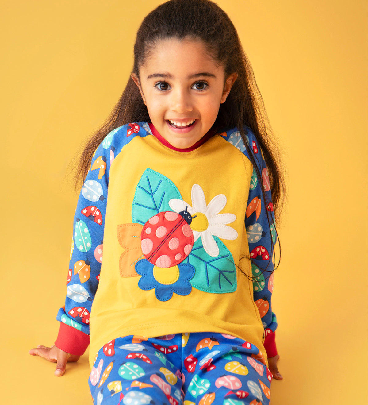 A child smiling and sitting on a orange surface with a light orange background. Wearing the mustard yellow top with a ladybird design on the front and blue sleeves with a repeating ladybird design and matching blue trousers 