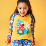 A child smiling and sitting on a orange surface with a light orange background. Wearing the mustard yellow top with a ladybird design on the front and blue sleeves with a repeating ladybird design and matching blue trousers 