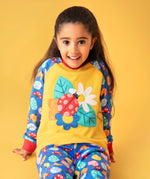 A child smiling and sitting on a orange surface with a light orange background. Wearing the mustard yellow top with a ladybird design on the front and blue sleeves with a repeating ladybird design and matching blue trousers 