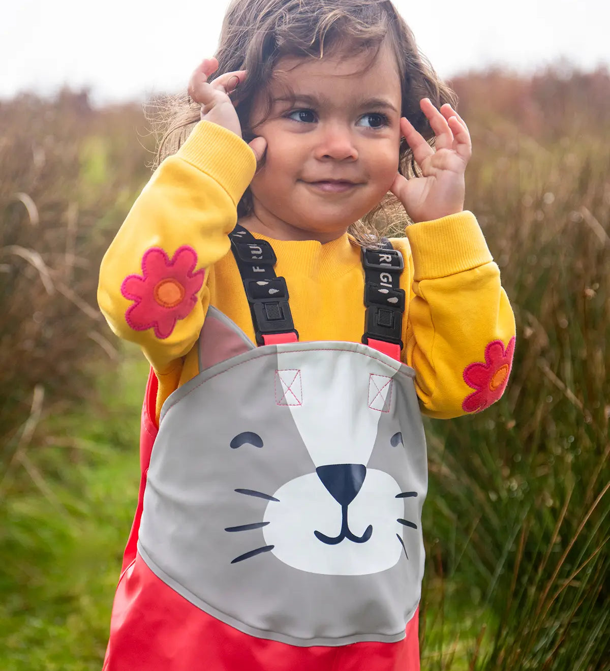 Small child wearing the Frugi character puddle buster trousers in rhubarb pink with a grey cat upper body panel