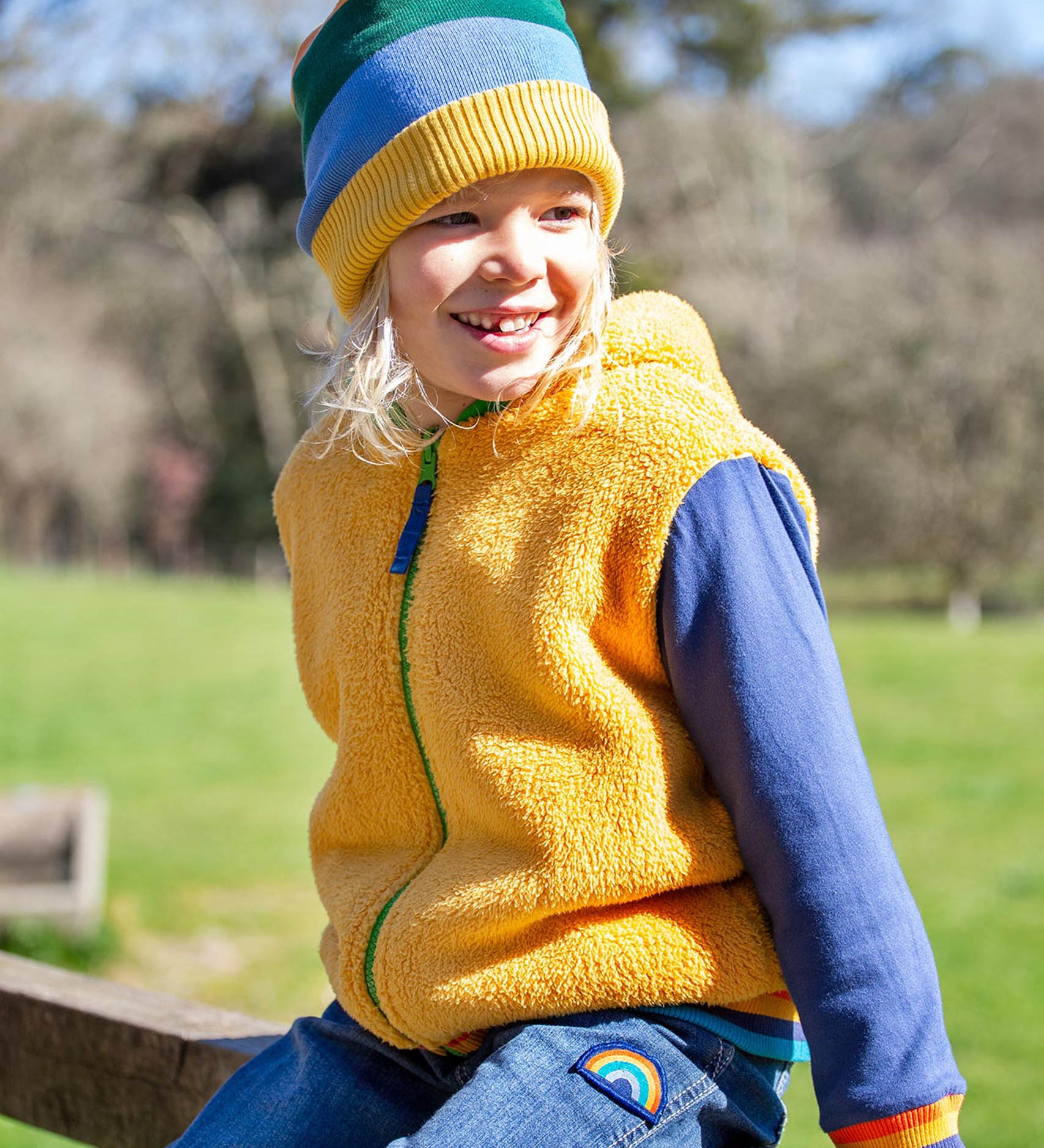 A child sitting outside and wearing the yellow ted fleece Frugi helston reversible hoodie.
