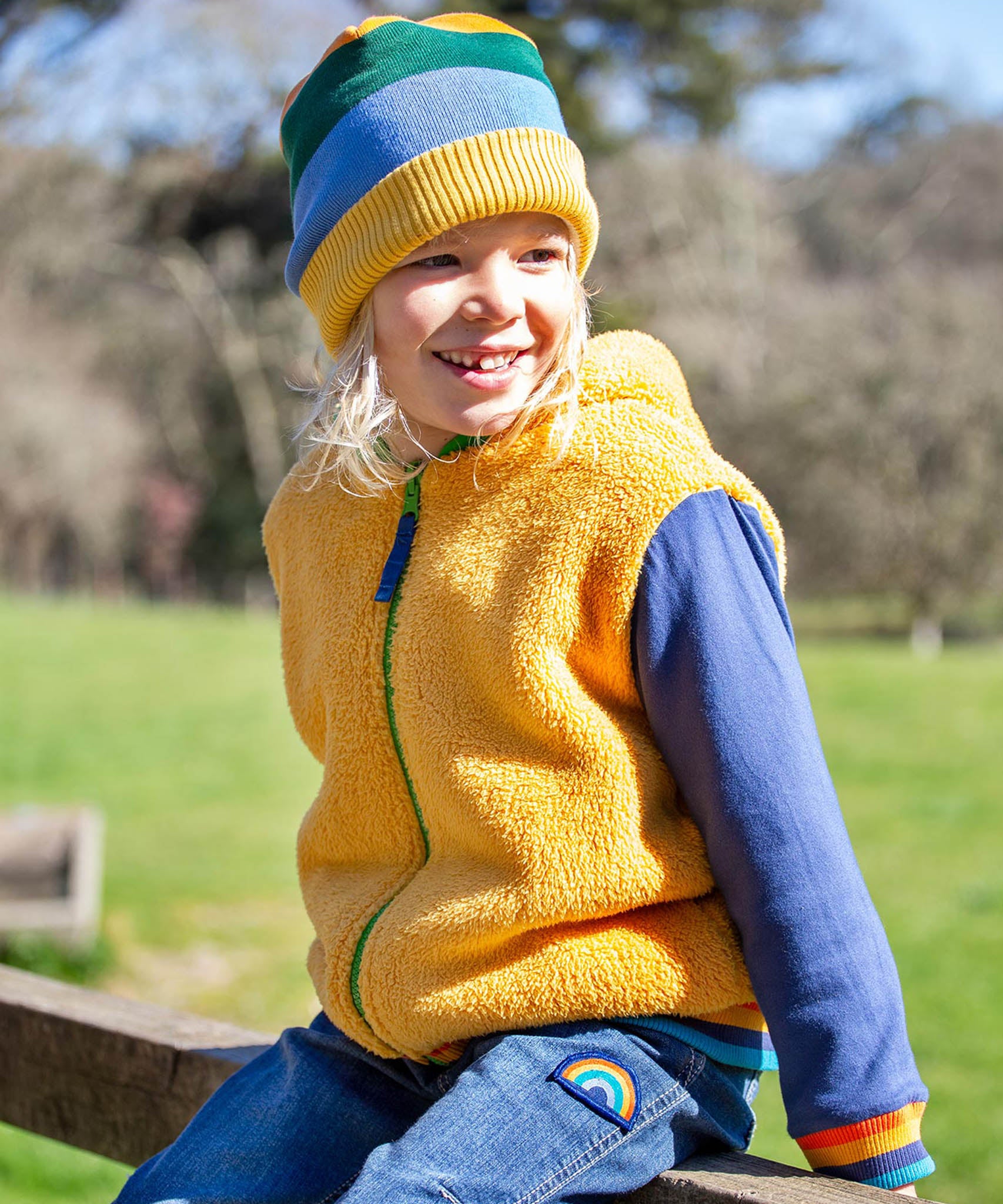 A child sitting outside and wearing the yellow ted fleece Frugi helston reversible hoodie.
