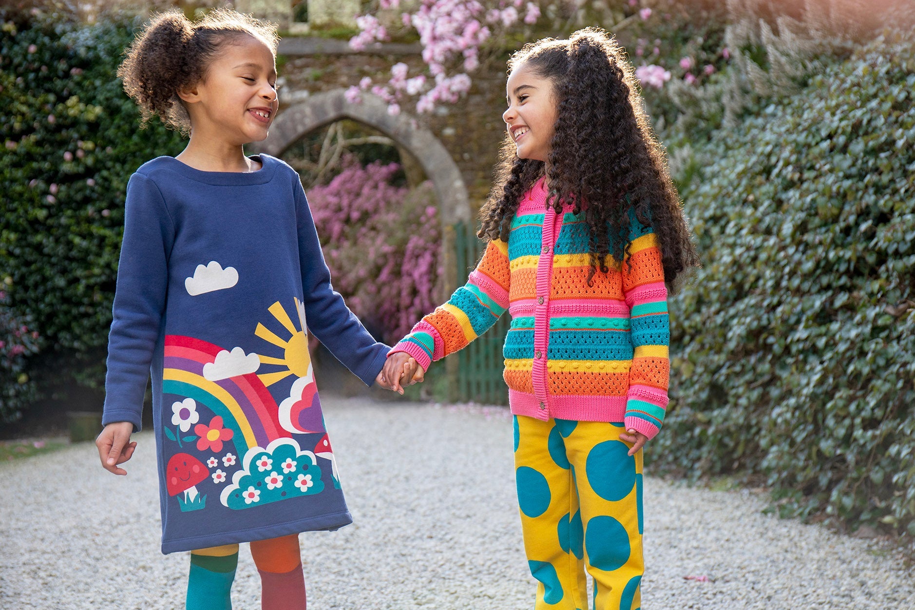 2 children standing outside in a garden. One child is wearing the Frugi Kari navy rainbow dress, other child is wearing the Frugi bright rainbow striped cardigan and super dot trousers available at Babipur.
