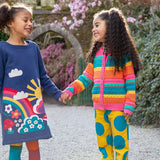 2 children standing outside in a garden. One child is wearing the Frugi Kari navy rainbow dress, other child is wearing the Frugi bright rainbow striped cardigan and super dot trousers available at Babipur.