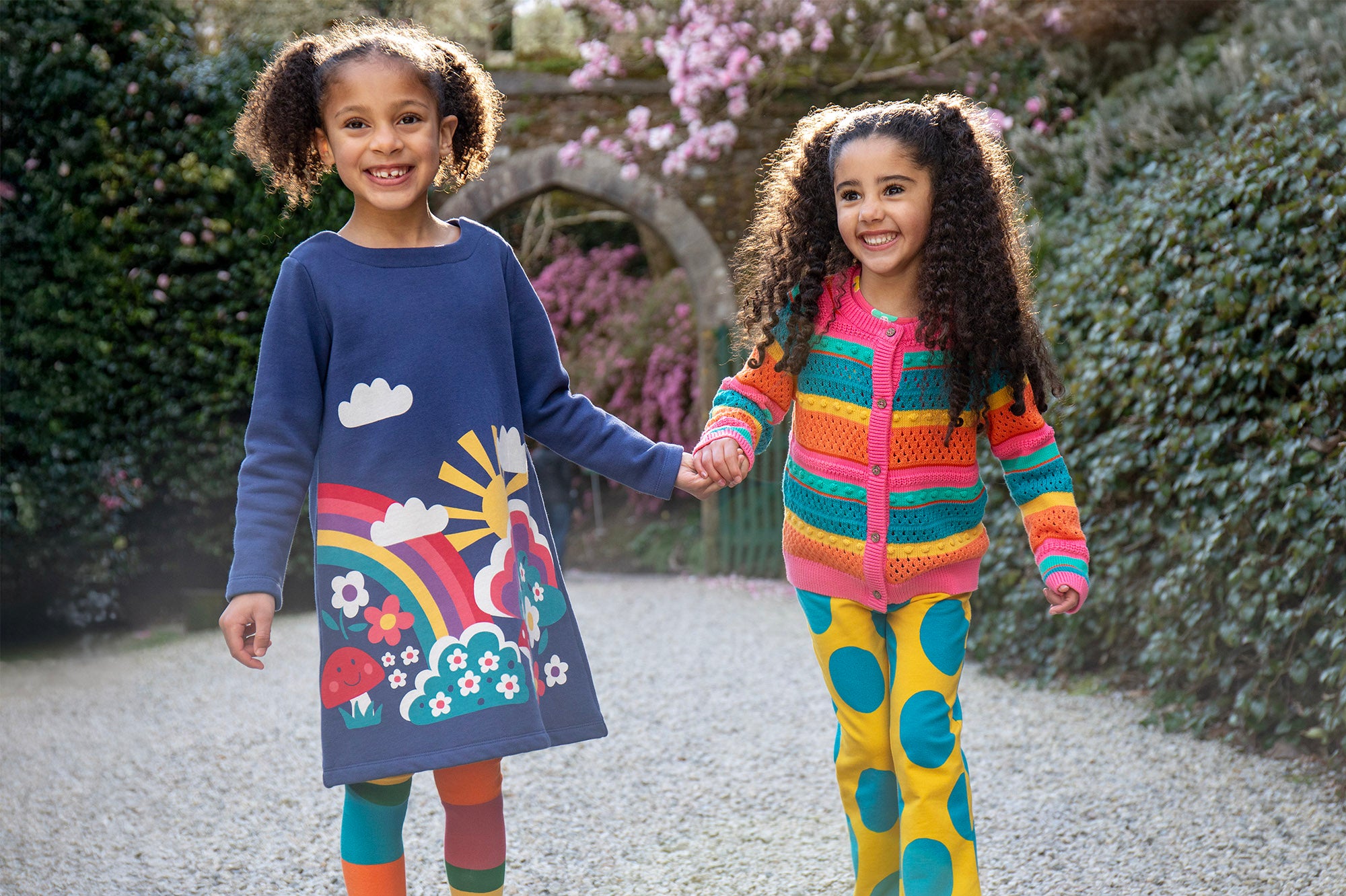 2 children holding hands in a garden setting. One child is wearing the Frugi Kari navy rainbow dress, the other child is wearing the Frugi bight rainbow striped cardigan and super dot trousers available at Babipur.