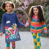 2 children holding hands in a garden setting. One child is wearing the Frugi Kari navy rainbow dress, the other child is wearing the Frugi bight rainbow striped cardigan and super dot trousers available at Babipur.
